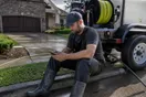A pressure washing operator sits on the curb next to his rig checking a slow-loading website on his phone after finishing a driveway job, in a natural documentary style.