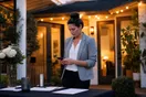 A real estate agent checks her phone at an empty open house sign-in table at dusk, in a natural documentary style.
