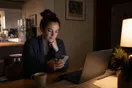 A woman sits at her kitchen table late at night with a laptop and a cup of tea, scrolling home listings on her phone before bed.