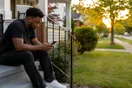 A person sits on a porch step scrolling a phone with a for-sale-style yard in soft focus behind them, waiting for a page to load, in warm evening light.
