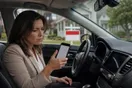 A real estate agent sits in her parked car outside a listing, checking a slow-loading page on her phone with a yard sign visible through the windshield, in a natural documentary style.