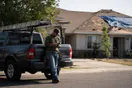 A roofer stands beside his parked pickup truck at the curb, checking his phone between storm-damage estimates on a quiet residential street.
