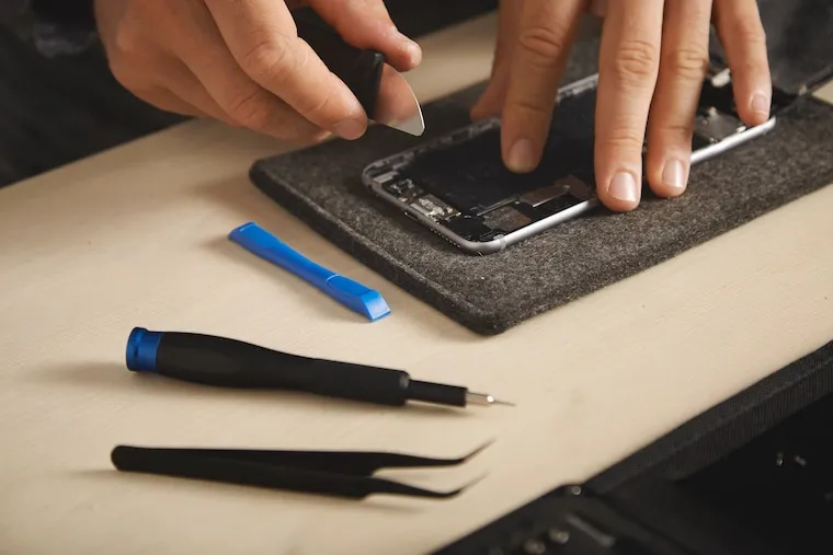 A phone repair workbench with precision screwdrivers, tweezers, a heat gun, and a microscope arranged on an anti-static mat, in a natural documentary style.
