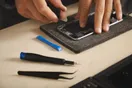 A phone repair workbench with precision screwdrivers, tweezers, a heat gun, and a microscope arranged on an anti-static mat, in a natural documentary style.