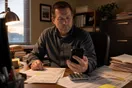 An accountant sits at a desk with tax documents spread out, checking a phone for a new client inquiry as afternoon light comes through the office window.