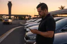 A car rental lot attendant checks his phone beside a row of parked rental sedans near an airport access road at dusk.