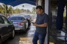 A car wash owner stands near the tunnel exit checking his phone as a line of cars waits under the entrance canopy.