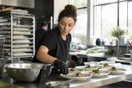 A caterer plates hors d'oeuvres in a bright commercial kitchen while checking her phone for a new inquiry propped against a mixing bowl.