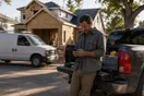 A general contractor stands beside a pickup truck outside a half-finished home addition, looking at his phone while a rival crew's van drives past.