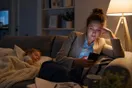 A mother sits on her couch late at night scrolling a daycare website on her phone while her toddler sleeps beside her.
