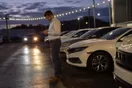 A car salesman stands on an evening dealership lot checking his phone beside a row of sedans, headlights from a passing car sweeping across the pavement.