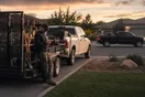 A landscaper stands beside his loaded trailer in a suburban driveway at dusk, checking his phone as a competitor's truck idles down the street.