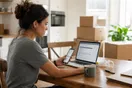 A woman sits at her kitchen table comparing moving company quotes on a laptop and phone side by side, moving boxes stacked behind her.