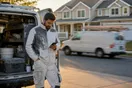 A painter stands at the tailgate of his van outside a suburban two-story house, checking his phone while a competitor's van passes on the street.