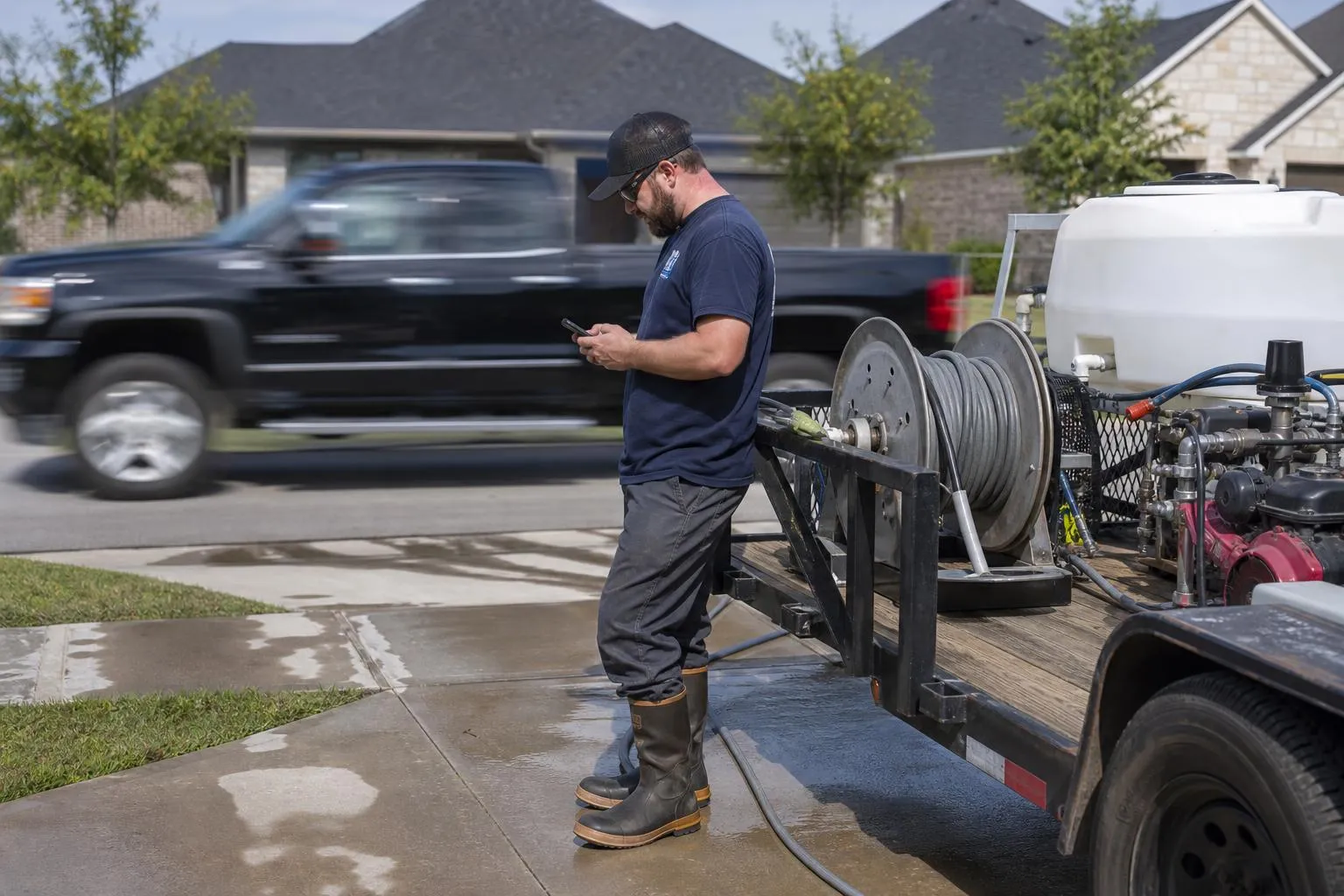 A pressure washing contractor stands beside his trailer-mounted rig in a suburban driveway, checking his phone while a competitor's truck passes behind him.