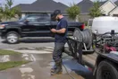 A pressure washing contractor stands beside his trailer-mounted rig in a suburban driveway, checking his phone while a competitor's truck passes behind him.