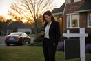 A real estate agent stands beside a for-sale sign in a suburban front yard at dusk, checking her phone while a rival agent's car pulls away from a nearby listing.
