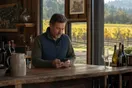 A vineyard owner stands at a tasting room counter checking a phone for new reservations while afternoon light falls across rows of vines outside.