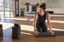 A yoga teacher rolls up a mat in a sunlit studio while glancing at a phone propped against a stack of blocks after morning class.