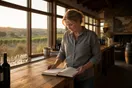 A winery owner stands alone in the tasting room at closing time looking at an empty reservation book, in a natural documentary style.