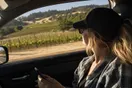 A passenger in a car holds a phone showing a vineyard map while rolling hills pass outside the window, photographed in a natural documentary style.