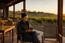 A winery owner checks her phone on the tasting room patio at golden hour, empty tables behind her waiting for a weekend crowd that hasn't arrived yet.