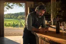 A vineyard owner leans on a tasting-room counter checking a slow-loading page on his phone as afternoon light falls across the rows outside, in a natural documentary style.