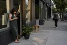 A studio owner stands outside her storefront checking her phone before opening, while a student walks past toward a competing studio down the block, in a natural documentary style.