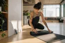 A yoga studio owner rolling out a mat in a sunlit studio while glancing at a phone propped against the wall before the first class.