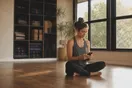 A yoga studio owner sits on the floor of an empty practice room scrolling her phone before the evening class, mats stacked against the wall in soft late light.