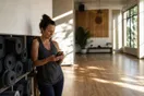 A yoga studio owner checks a phone in a quiet sunlit studio with rolled mats stacked along the wall, natural documentary light.